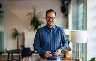 Middle-aged man in blue button up and glasses standing next to window and smiling
